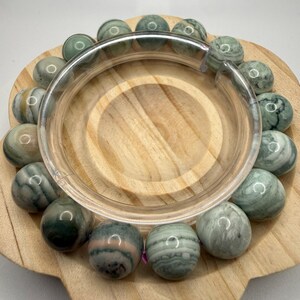 May include: A circular bracelet composed of smooth, spherical beads in shades of sage green, white, and grey with subtle marbled patterns. The beads are arranged in a continuous loop, showcasing natural variations in color and texture throughout.
