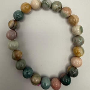 May include: A circular stretch bracelet composed of smooth, round beads in a variety of colors including shades of sage green, soft pink, warm tan, and cream. The beads feature natural marbling and variations in tone throughout the surface.