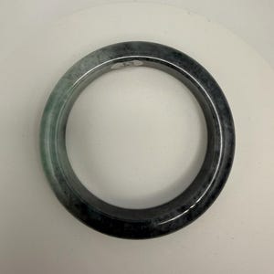 May include: A round jade bangle bracelet. The bracelet is a mix of dark green and black. The jade is polished and has a smooth surface. The bracelet is a piece of jewelry.