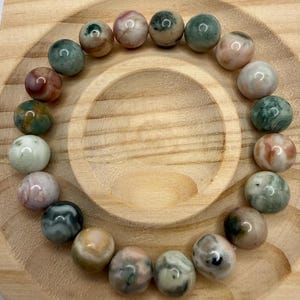 May include: A circular bracelet composed of smooth, polished spherical beads in various shades of green, pink, cream, and brown. The beads feature natural swirling patterns and inclusions, arranged in a continuous loop on a light wooden surface.