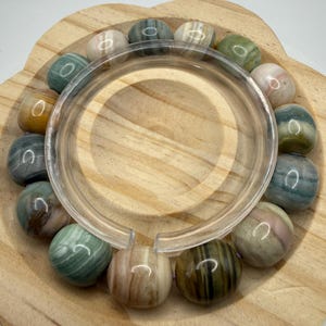 May include: A circular bracelet composed of smooth, polished, multi-colored stone beads in shades of green, brown, gray, and cream. The beads feature natural banded patterns and are strung together, encircling a clear, C-shaped plastic display stand.