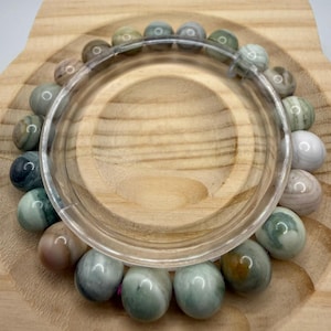 May include: A circular bracelet composed of smooth, polished spherical beads in varying shades of sage green, soft pink, cream, and muted gray. The beads are strung together in a continuous loop, displaying natural marbled patterns and glossy finishes.