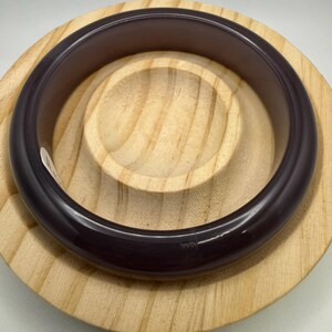 May include: A dark purple bangle bracelet with a smooth, glossy finish. The bracelet is circular and appears to be made of a solid material. It rests on a light-colored wooden, circular display stand.
