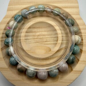 May include: A circular bracelet composed of smooth, round beads in varying shades of sage green, dusty rose, and cream. The beads are strung together and arranged in a continuous loop, displaying natural color variations and subtle surface textures.