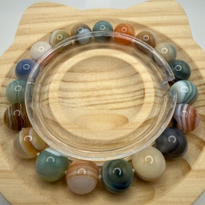 May include: A circular bracelet composed of smooth, polished, multi-colored agate beads in shades of blue, green, brown, orange, white, and gray. Each bead features distinct natural banding patterns, and the bracelet is strung on a flexible cord.