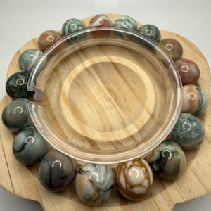 May include: A circular bracelet composed of polished, multi-colored stone beads in shades of sage green, cream, terracotta, and charcoal gray. The beads feature natural swirling patterns and are arranged in a continuous loop around a clear plastic ring.