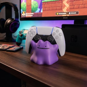 Könnte beinhalten: Ein violetter Controller-Ständer in Ditto-Form, der einen weiß-schwarzen PlayStation 5-Controller hält. Eine schwarze Nintendo Switch-Konsole ist zu sehen, zusammen mit einem Gaming-Monitor und Kopfhörern.