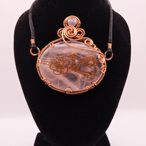 Può includere: Collana con pendente artigianale in filo di rame. Il pendente presenta una grande pietra ovale traslucida con un disegno floreale. La pietra è racchiusa in filo di rame e pende da un cordino nero.