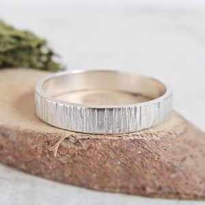May include: A silver ring with a textured bark-like design. The ring is sitting on a piece of wood.