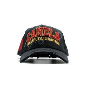 Può includere: Cappellino da baseball nero con la scritta "CANELO" ricamata in rosso e oro. Il testo "UNDISPUTED CHAMPION" è ricamato sotto. Il cappellino ha una visiera usurata e una toppa rossa con paillettes sul lato.