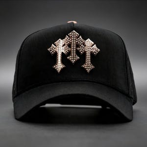 May include: A black baseball cap featuring a prominent metallic rose gold emblem on the front. The emblem consists of three interconnected cross shapes, each densely encrusted with small, sparkling clear stones that catch the light.