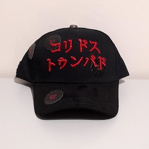 May include: Black baseball cap with red Japanese text and floral accents. The cap has embroidered Japanese characters on the front and small floral designs on the sides. A stylish accessory for casual wear.