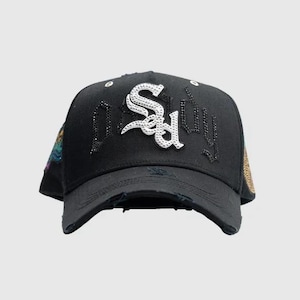 Puede incluir: Gorra de béisbol negra con borde desgastado. La gorra presenta la palabra "Sadboy" en una fuente blanca y adornada en la parte delantera. La gorra tiene un parche decorativo en el lateral y está sobre un fondo liso.