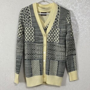 May include: A long-sleeve V-neck cardigan featuring a patchwork pattern of black, white, and gray geometric designs. The garment has a solid pale yellow trim along the button placket, neckline, and cuffs, with five matching pale yellow buttons.