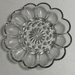 May include: Clear glass serving dish with a scalloped edge. The dish has a central starburst design and individual compartments around the edge. The glass is transparent, allowing light to pass through, and is suitable for serving appetizers or small portions.