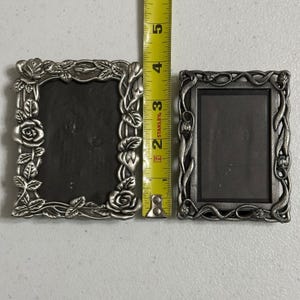 May include: Two rectangular silver-toned metal picture frames with raised floral relief designs. The left frame features rose motifs, while the right frame has vine patterns. A yellow Stanley measuring tape is placed between them, showing a height of 5 inches.