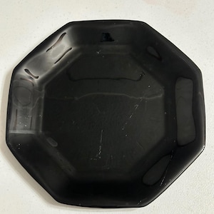 May include: A black, octagonal plate with a glossy finish. The plate has a slightly raised edge and a smooth surface. The plate is empty and appears to be made of a durable material. The plate is suitable for serving food.
