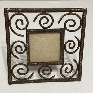 May include: A square metal picture frame with a dark bronze finish and a textured, cross-hatched border. The frame features four decorative metal spirals positioned at each corner, surrounding a central square opening with a plain beige insert.