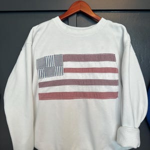 May include: White sweatshirt with a sewn-on American flag design. The flag features a blue and white checkered square in the upper left corner, with red and white striped sections. The sweatshirt has a crew neck and cuffed sleeves.
