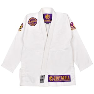 May include: White martial arts uniform with purple and gold embroidered logos. The uniform features the text "SHOYOROLL" on the chest, sleeves, and lower front. The uniform is designed for martial arts training and competition.