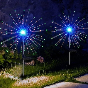 Può includere: Due luci da giardino a energia solare con design a fuoco d'artificio. Ogni luce ha un LED blu centrale circondato da steli radianti con luci multicolori. Le luci sono montate su pali neri con pannelli solari, su uno sfondo scuro.
