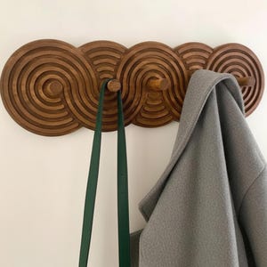 Può includere: Un attaccapanni da parete in legno dal design unico. L'attaccapanni è realizzato in legno marrone scuro e presenta una serie di cerchi concentrici. Una borsa verde e un cappotto grigio sono appesi all'attaccapanni, mostrando la sua funzionalità e il suo stile.