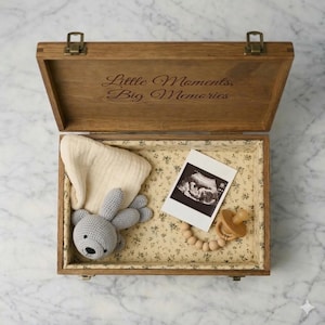 May include: A wooden keepsake box, engraved with "Little Moments, Big Memories", is open to reveal a gray crocheted bunny, a cream muslin cloth, an ultrasound scan, and a wooden dummy with a beaded chain, all on floral fabric.