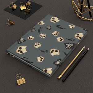 Puede incluir: Un diario gris con un patrón repetido de calaveras beige y formas negras en forma de garra. Lápices dorados y negros, junto con clips dorados, están dispuestos alrededor del diario sobre una superficie gris oscuro.