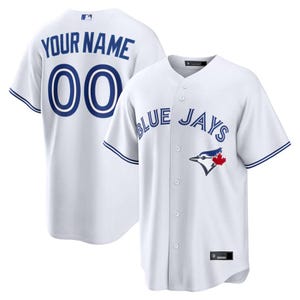 Puede incluir: Camiseta de béisbol blanca con detalles azules. La parte delantera muestra "BLUE JAYS" y un logotipo de un arrendajo azul con una hoja de arce roja. La parte trasera tiene "YOUR NAME" sobre el número "00".