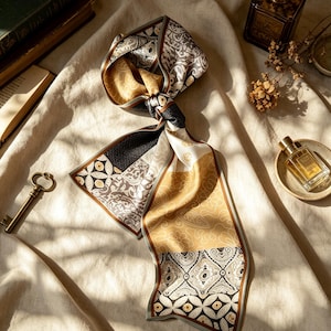 May include: A long, narrow scarf featuring a patchwork design of various patterns in shades of gold, black, white, and grey. The scarf is tied in a knot at the center, displaying geometric, floral, and abstract motifs across its rectangular fabric panels.