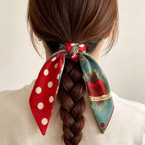 May include: A hair scrunchie featuring a long, tied ribbon design. One side of the ribbon is red with white polka dots, while the other side is sage green with a graphic illustration of a red heart and the text Te Amo Mucho.