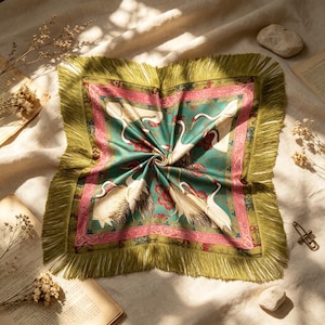 May include: A square scarf featuring a teal green background with white crane illustrations, red floral accents, and a pink decorative border. The scarf is finished with long, olive green fringe trim along all four edges of the fabric.