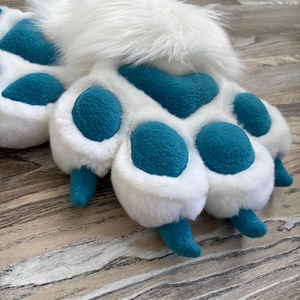 May include: A pair of plush animal paw gloves featuring long white faux fur on the back and soft teal fabric on the palm. The design includes four rounded teal paw pads and four pointed teal claws extending from the white fur-covered fingers.