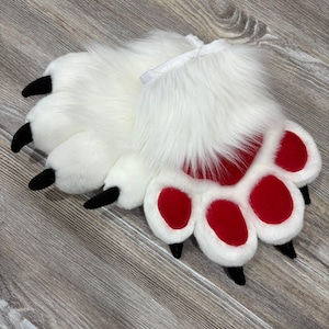 May include: A pair of white faux fur paw slippers with red paw pads and black claws. The slippers have a fluffy texture and are designed to resemble animal paws. The cuffs are white.