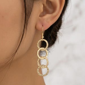 May include: Gold-toned dangle earrings featuring four textured, interlocking circles. The earrings have a hook closure and are shown worn on an ear. The jewelry has a delicate, feminine design.
