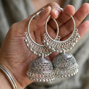 May include: A pair of silver-colored hoop earrings with a bell-shaped design. The earrings feature intricate detailing and small dangling beads along the bottom and around the hoop. The earrings are held in a hand.