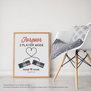 May include: A framed print with a white background and red text that reads "Forever in 2 Player Mode". Two retro video game controllers are connected by a red heart-shaped cord. The text "NAME NAME May 27th, 2016" is below the controllers.