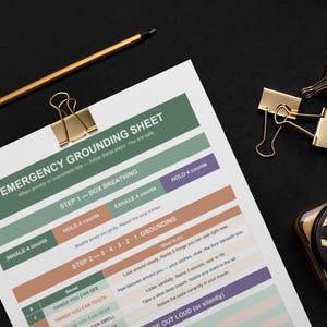 May include: A printed "Emergency Grounding Sheet" with instructions for managing anxiety, featuring steps for box breathing and grounding techniques. Includes a gold pencil, paper clips, and a decorative container on a black surface.