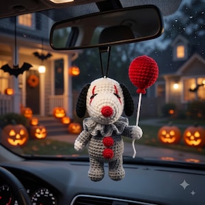 Könnte beinhalten: Eine gehäkelte Hundefigur, die an einem Rückspiegel hängt, gestaltet als Clown mit weißem Kopf, schwarzen Ohren, roter Nase und roten Augenmarkierungen. Sie trägt einen grauen Rüschenkragen und einen grauen Körper mit zwei roten Knöpfen.