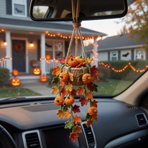 Könnte beinhalten: Ein gehäkeltes Hängepflanzen-Ornament mit einem kleinen beigen Korb, gefüllt mit Miniatur-Kürbissen in Orange und herbstlichen Blättern in Rot, Orange, Gelb und Grün, das an einer beigen Schnur am Rückspiegel eines Fahrzeugs befestigt ist.