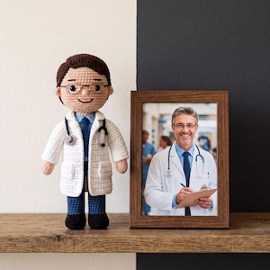 May include: A crocheted doll of a doctor with brown hair, glasses, a white lab coat, a blue tie, and a stethoscope, standing next to a wooden picture frame containing a photograph of a doctor in a white coat with a stethoscope and a clipboard.