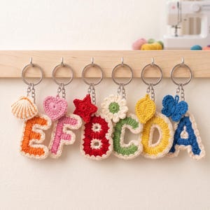 May include: Six crochet letter keychains hanging on a wooden rack. Each letter features a cream-coloured border and a unique charm: a seashell on orange E, a heart on pink F, a star on red B, a flower on green C, a leaf on yellow D, and a butterfly on blue A.