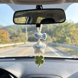 May include: A crochet car hanging ornament featuring a light blue fish, a cream-colored star with a small blue bow, and a green four-leaf clover, all connected by a cream-colored yarn loop for suspension from a rearview mirror.