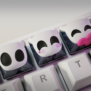 May include: Close-up of three custom keycaps with white faces and black eyes, each with a unique expression. One keycap has a pink tongue. The keycaps are on a white keyboard with gray and black accents.