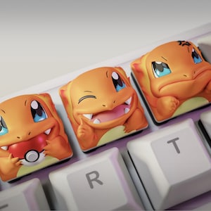 May include: Three orange Charmander keycaps with varied expressions, including a happy, winking, and sad face. The keycaps are on a white keyboard, with the letters "R" and "T" visible.