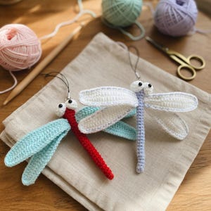 May include: Two handmade crochet dragonfly ornaments. One has a red body and blue wings, the other has a light purple body and white wings. The wings are detailed with white crochet. The ornaments are on a beige linen surface.