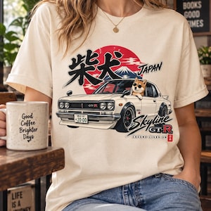 Peut inclure: Un t-shirt crème arborant un graphique d'une Nissan Skyline GT-R blanche avec un Shiba Inu portant des lunettes de soleil au volant. Le motif inclut un soleil rouge, le mont Fuji, des caractères japonais et le texte Skyline GT-R Legend Lives On.