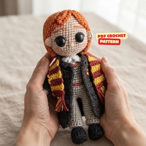 May include: A crocheted doll with orange hair, large black eyes, and a light skin tone. The doll wears a gray sweater, a dark gray robe, and a yellow and maroon striped scarf. A red rectangular label with the text PDF CROCHET PATTERN is visible.