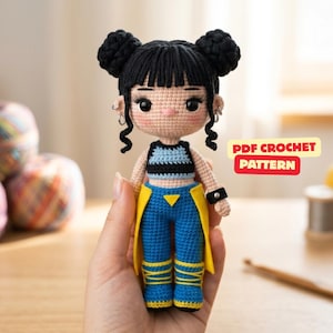 Può includere: Una bambola lavorata all'uncinetto a mano con capelli neri raccolti in due chignon e frangia. La bambola indossa un top corto a righe nere e blu, pantaloni blu con dettagli gialli e piccoli orecchini a cerchio argentati. Scritta: PDF CROCHET PATTERN.