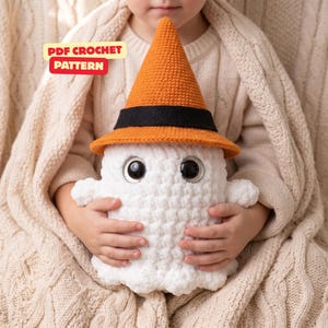 May include: A white crocheted ghost plush toy with large black eyes, wearing an orange pointed hat with a black band. The image includes the text PDF CROCHET PATTERN in a yellow and red box. The ghost has a soft, textured yarn finish.